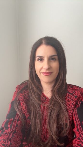 A photo of Dr Randa Abdel-Fattah. She has long brown hair parted in the middle and brown eyes. She is wearing a red top and muted red lipstick, and smiling into the camera.