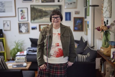 Pip Adam standing in a living room room. She has a jacket and glasses on and is smiling at the camera. The wall behind her is covered in framed pictures and art.