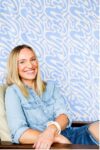 Broke Crawford sitting on a hair in front of a blue wallpapered wall. She is smiling at the camera.