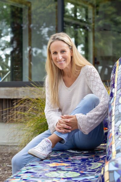 Shara Curlett sitting on a blue couch outdoors with her leg up and arms wrapped around her shin.
