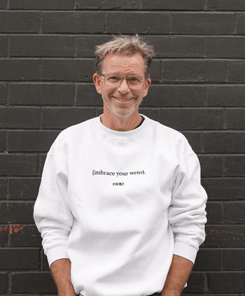 A photo of Ben Crowe. He stands in front of a black brick wall. He wears glasses and a white jumper.