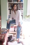 A photo of Lisa Wilkinson in front of a bookshelf. She wears a white shirt and jeans, and is resting one foot on a ledge.