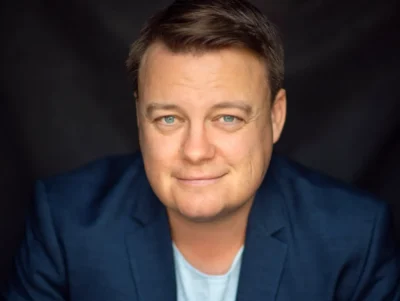 A photo of Luke Carman. A man is smiling at the camera and wears a navy blazer over a blue shirt.