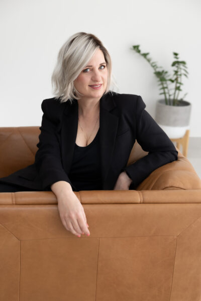 A photo of Marny Lishman. She is wearing black and sits on a brown couch. A potted plant is in the background.