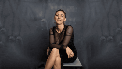 A photo of Mykaela Saunders sitting before a black background. Saunders has short hair and is wearing black.