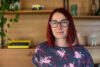 A photo of Stef Gemmill. Gemmill wears glasses and has red hair. She stands in front of a bookcase.