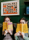 Marina and Xuan holding books over their mouths. A sign in the back reads "decolonise your bookshelf".